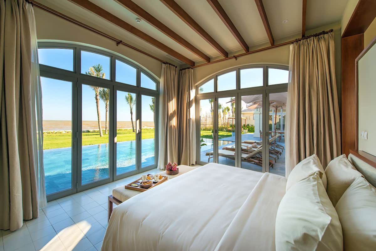 A spacious bedroom with a large bed, a breakfast tray, and a view of a swimming pool and palm trees.