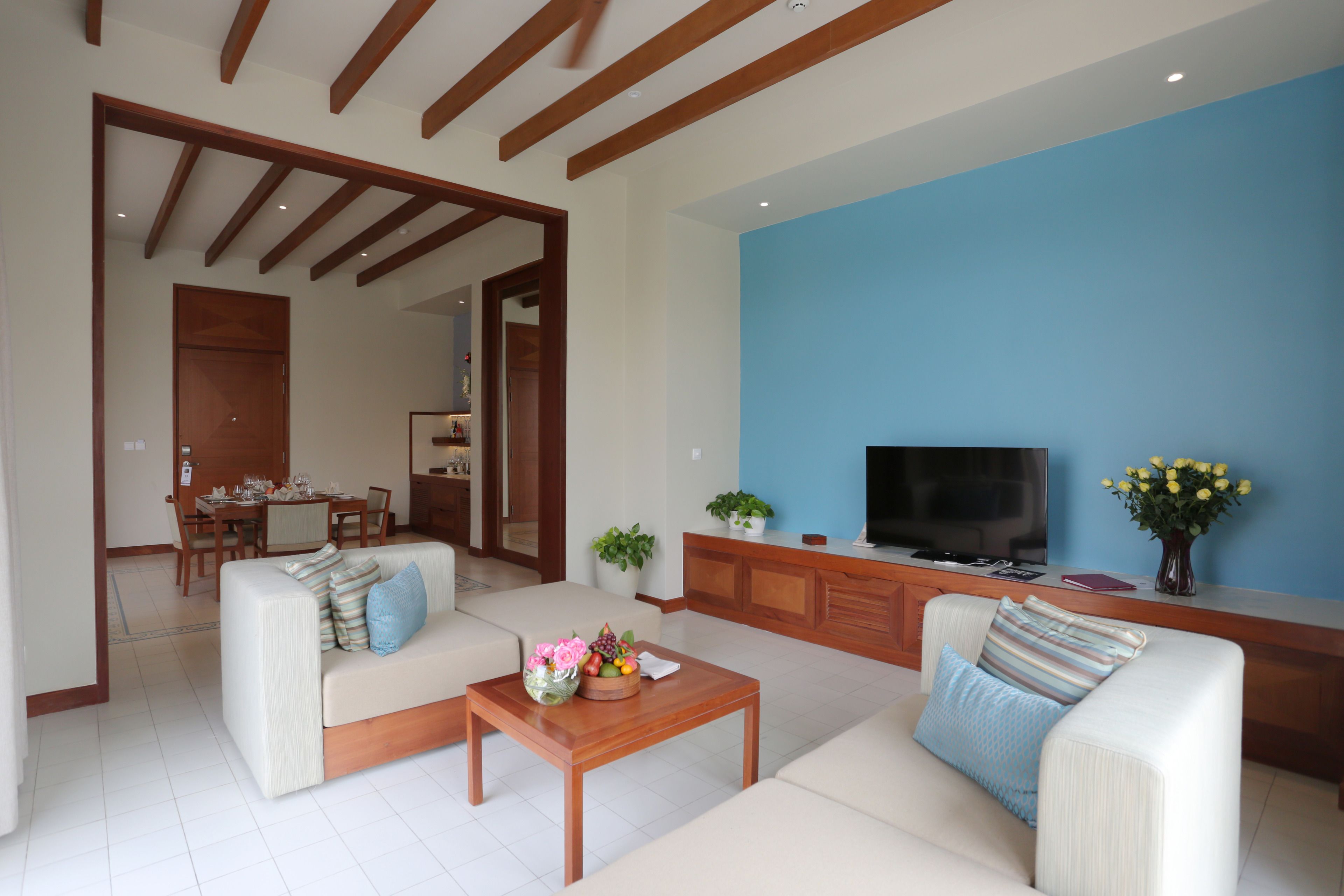 A modern living room with a sofa set, a coffee table with fruit and flowers, a TV, and a dining area in the background.