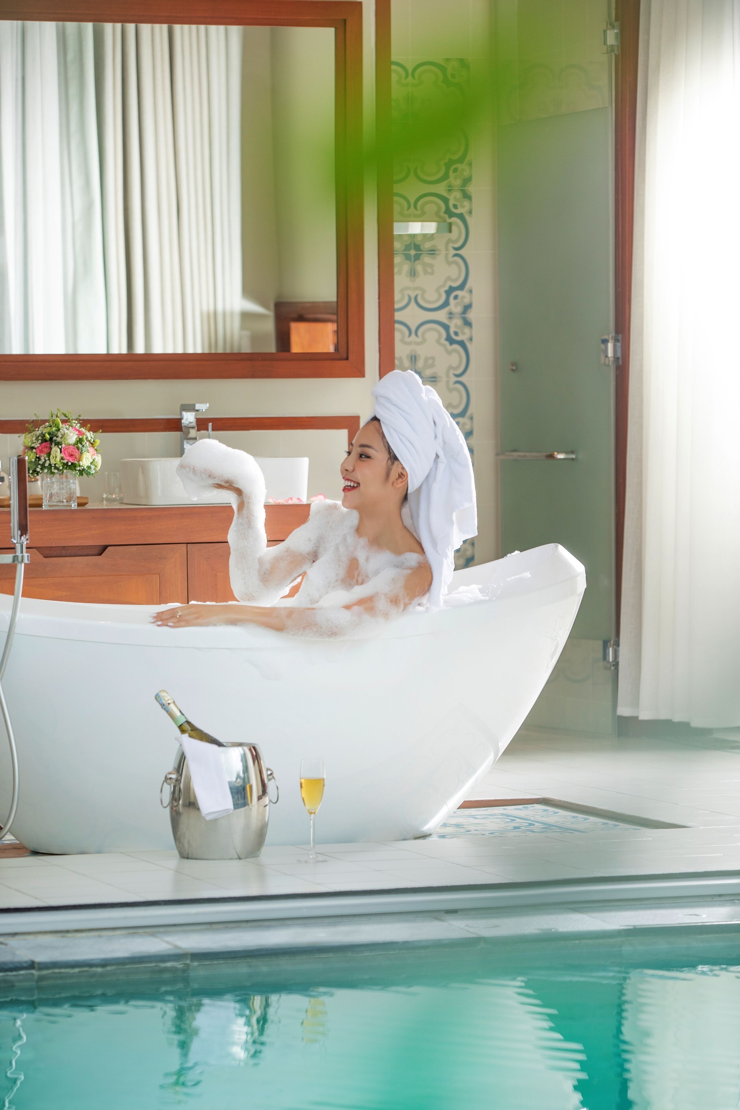 A woman in a white bath tub with a towel on her head, a champagne bottle, and a glass of champagne on the floor beside the tub.