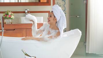 A woman in a white bath tub with a towel on her head, a champagne bottle, and a glass of champagne on the floor beside the tub.