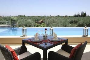Mesa de comedor dispuesta con servilletas blancas, cubiertos y vasos, con vistas a una piscina bajo un cielo azul despejado y vegetación al fondo.