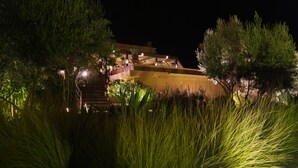 Un edificio de varios pisos con un diseño arquitectónico moderno, rodeado de abundante vegetación e iluminado con luz ambiental.