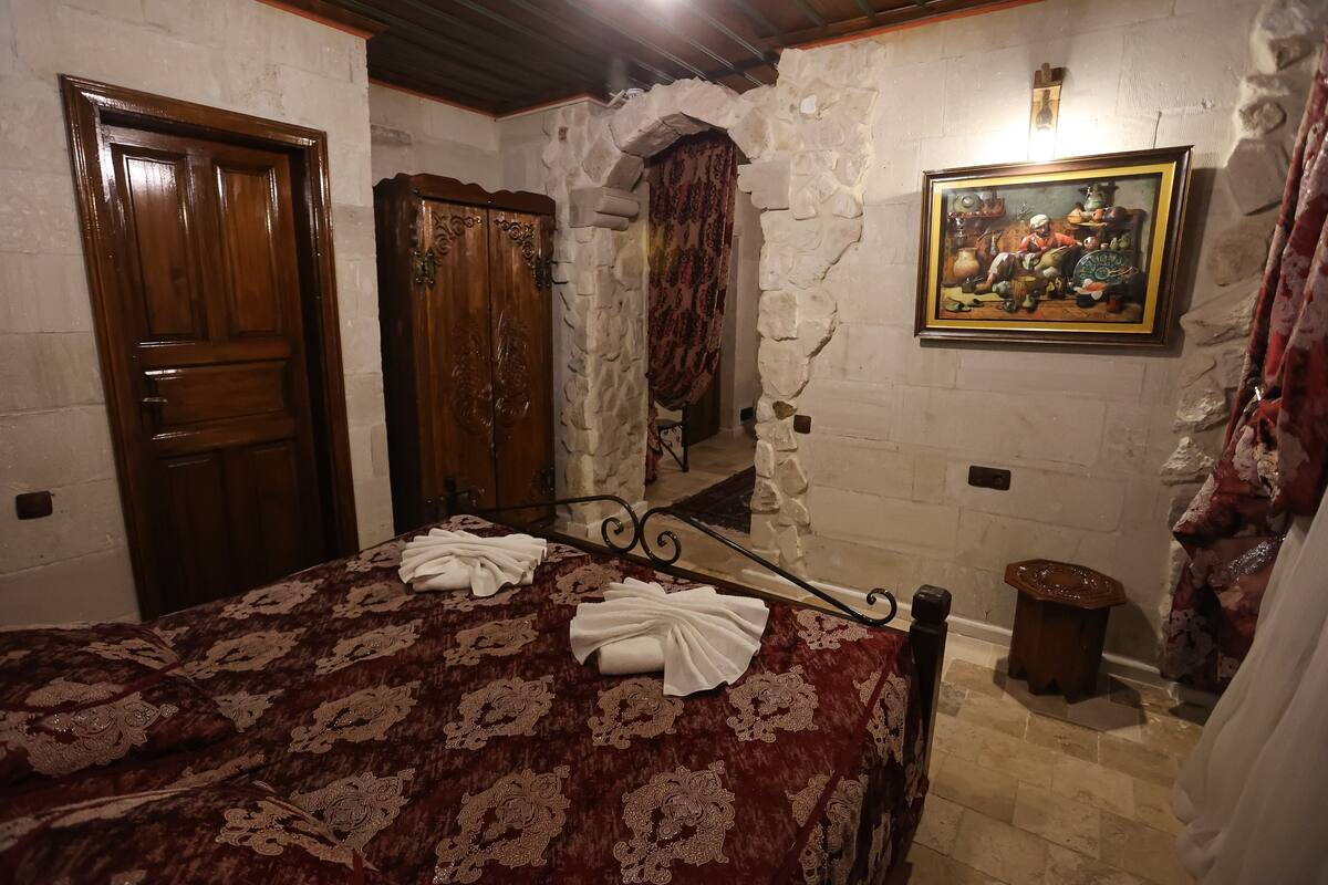 Una habitación con una cama, un armario de madera, una pared de piedra, un cuadro y un taburete pequeño.