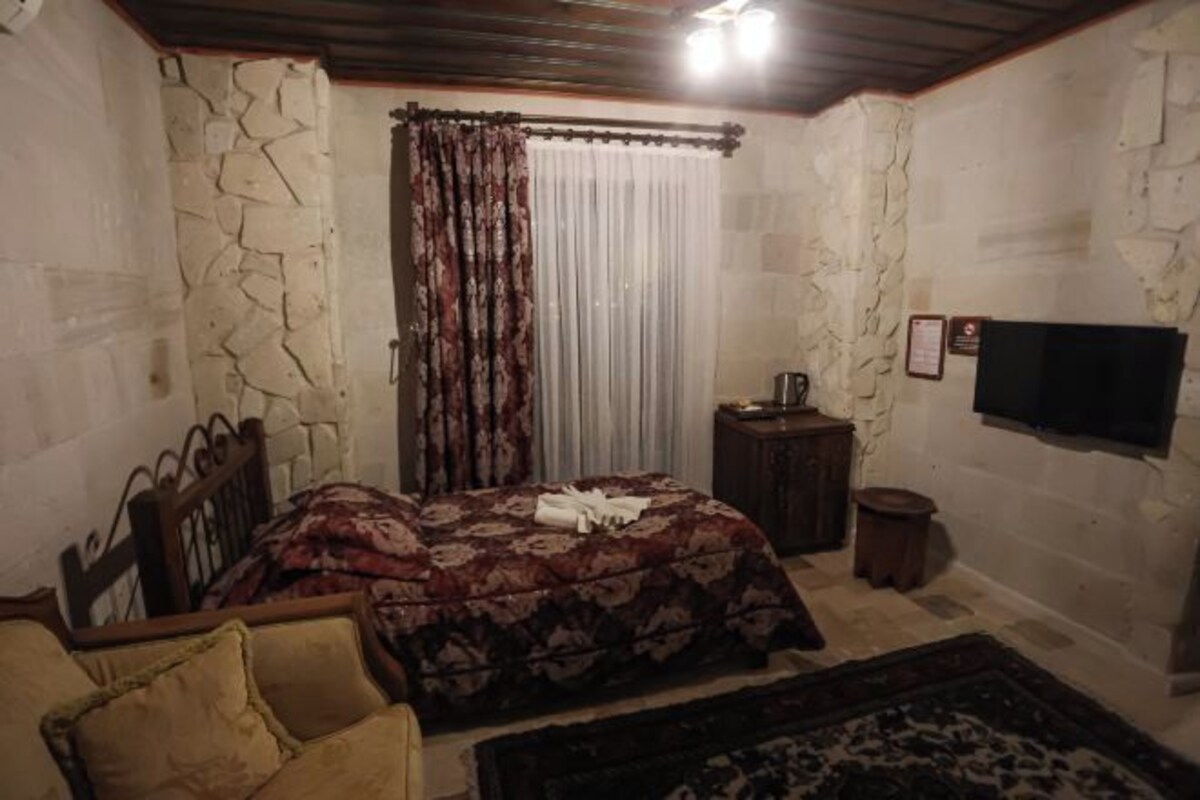 A bedroom with a bed, a wooden headboard, a small bedside table, a television mounted on the wall, and a window with curtains.
