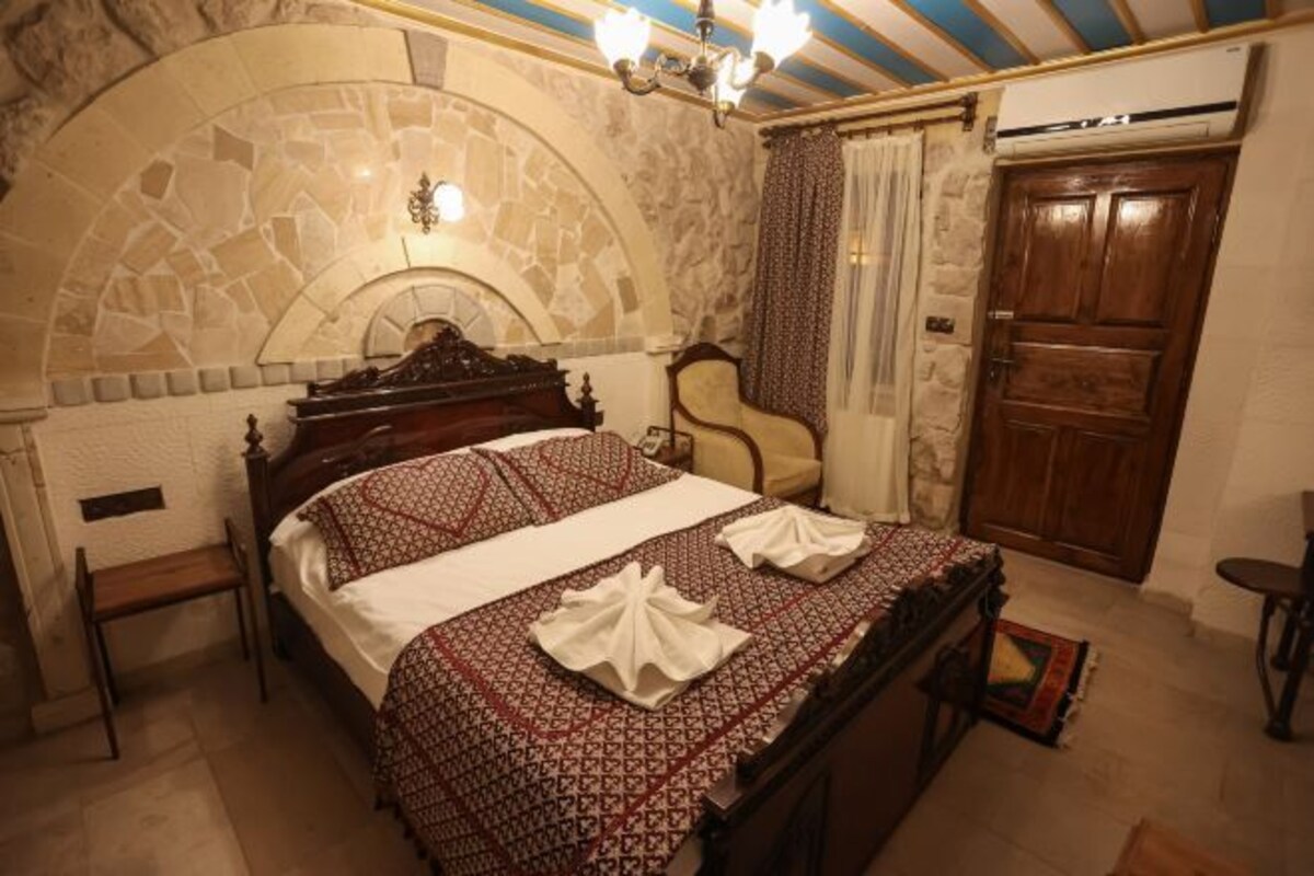 A bedroom with a stone arch, a bed with patterned bedding, a wooden wardrobe, and a chandelier.
