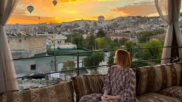 A woman sitting on a patterned sofa by a window overlooking a cityscape with hot air balloons during sunset.