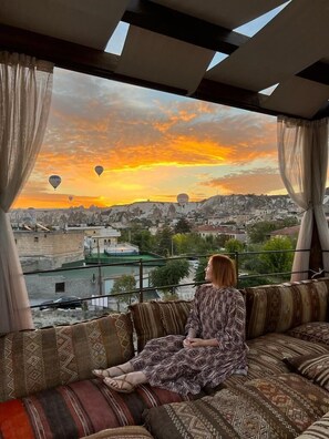 A woman sitting on a patterned sofa by a window overlooking a cityscape with hot air balloons during sunset. - Alaturca House (Nevsehir)