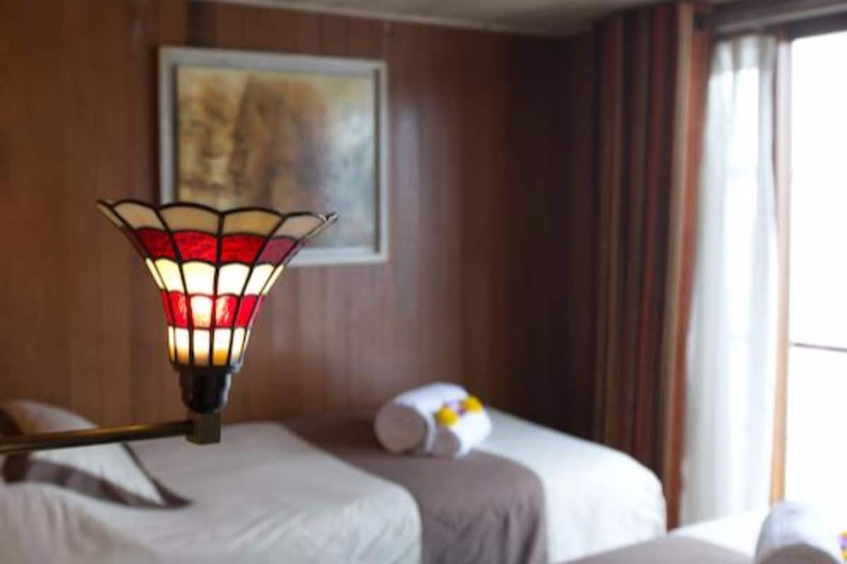 A hotel room with two beds, a wooden ceiling, a painting on the wall, and a window with curtains.