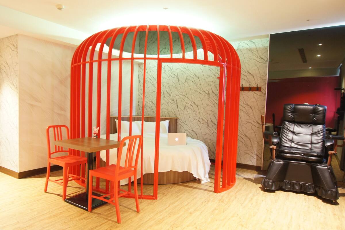 A hotel room with a bed, a desk with two chairs, a red partition, and a massage chair.