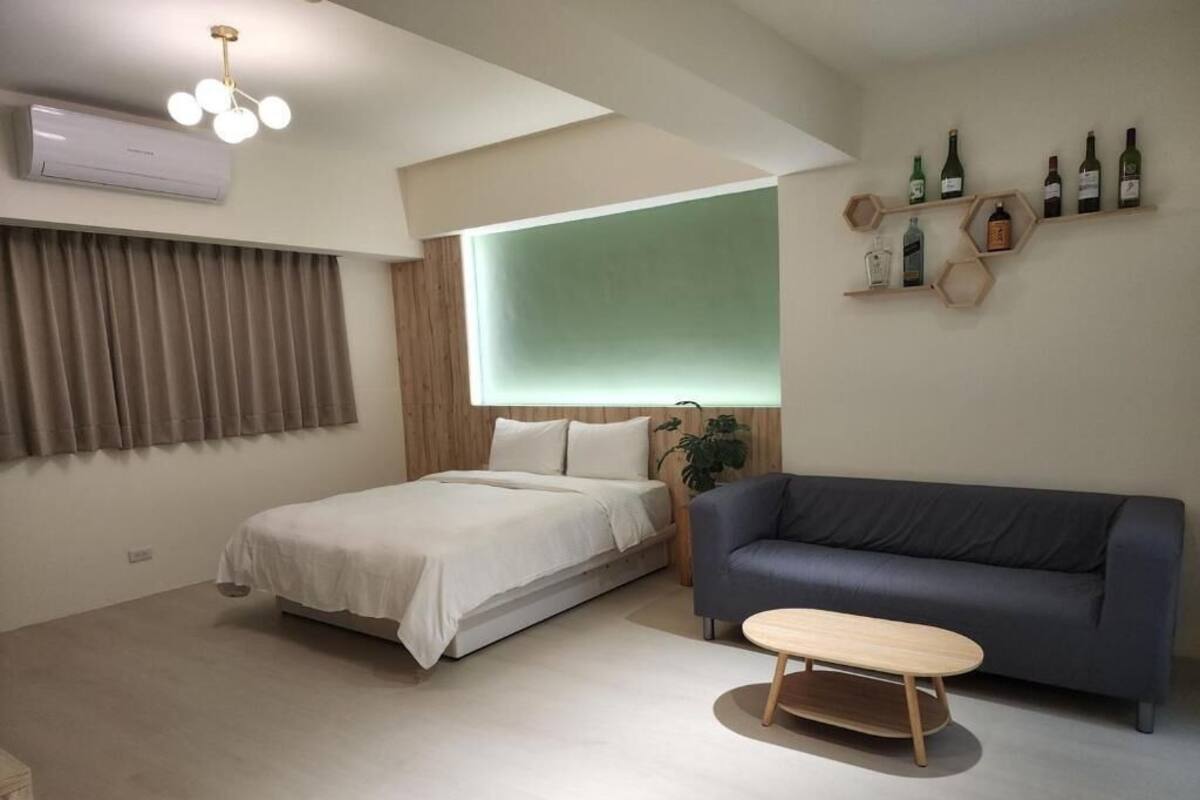 A modern room with a bed, a sofa, a small table, and a wall-mounted shelf with bottles.