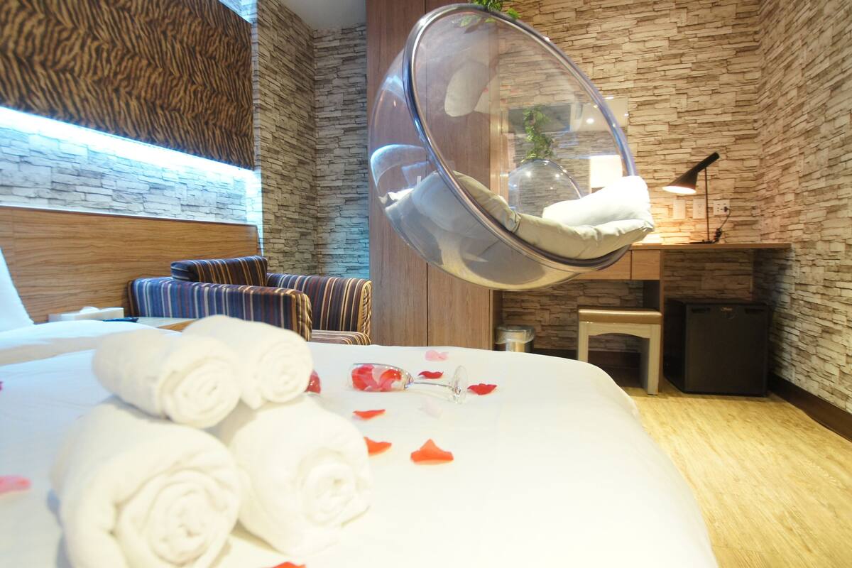 A hotel room with a bed, a hanging chair, and a stone wall.