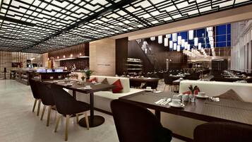 A modern restaurant with a geometric ceiling design, a central seating area with a sofa and dining tables, and a bar area with a staircase.