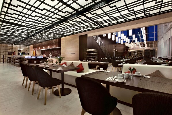 A modern restaurant with a geometric ceiling design, a central seating area with a sofa and dining tables, and a bar area with a staircase.