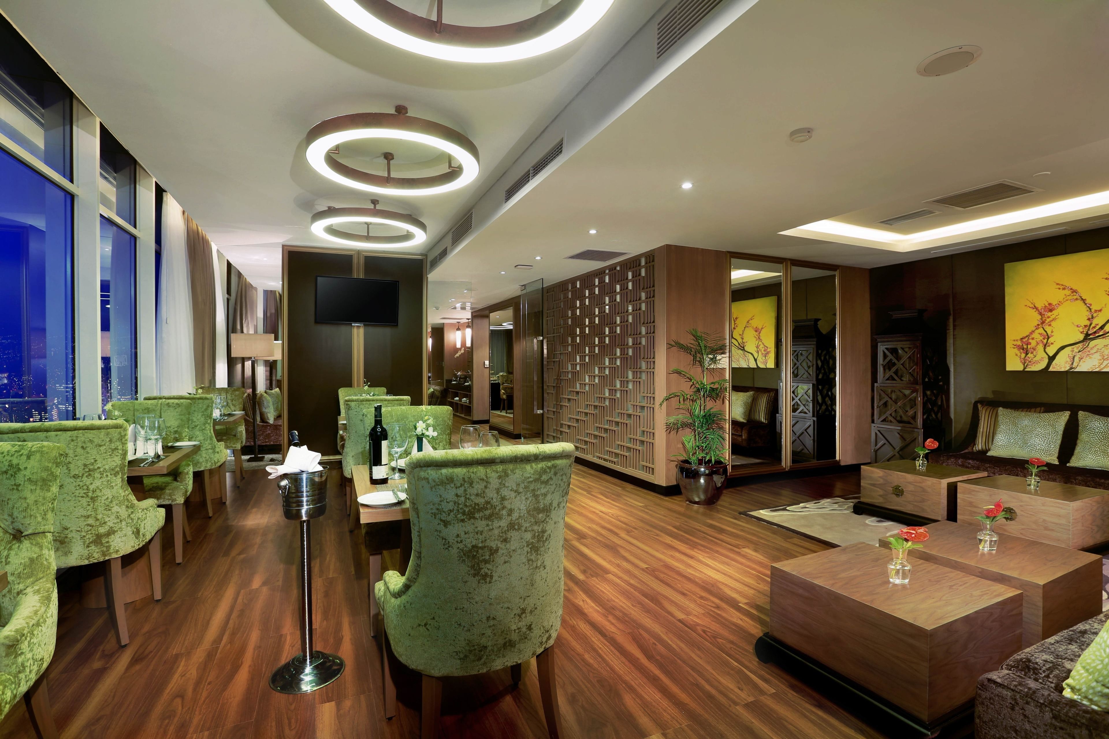 A modern hotel lobby with green upholstered seating, wooden flooring, and a large TV screen.