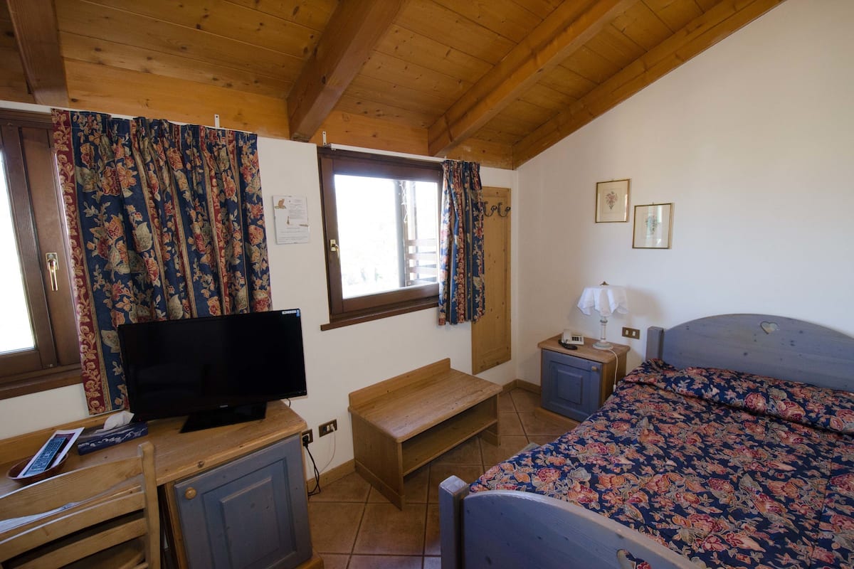 A bedroom with a bed, a desk with a television, a window with curtains, a bedside table, and a wooden ceiling.