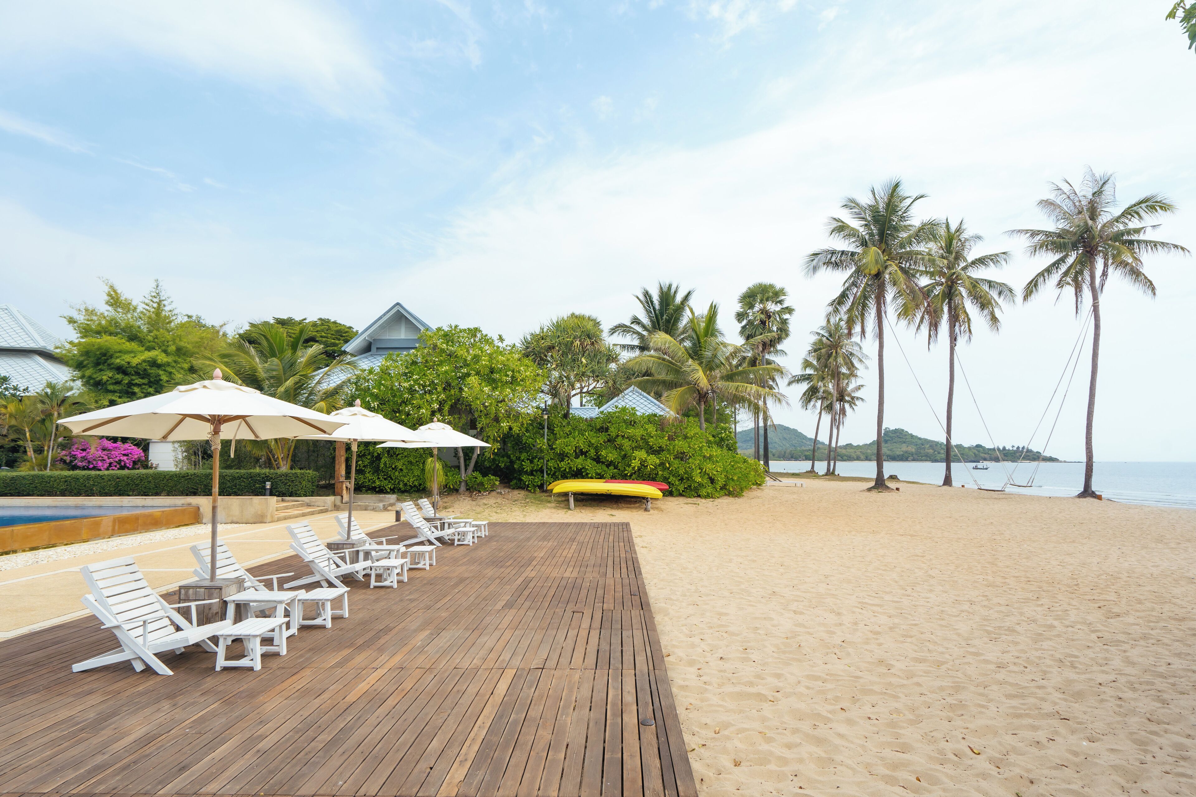 Una terraza de madera con sillones blancos y sombrillas, una piscina y una playa con palmeras y vistas al mar.