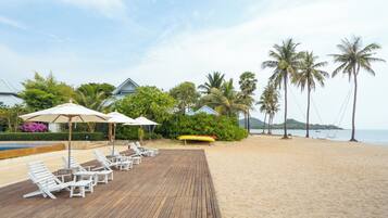 Una terraza de madera con sillones blancos y sombrillas, una piscina y una playa con palmeras y vistas al mar.