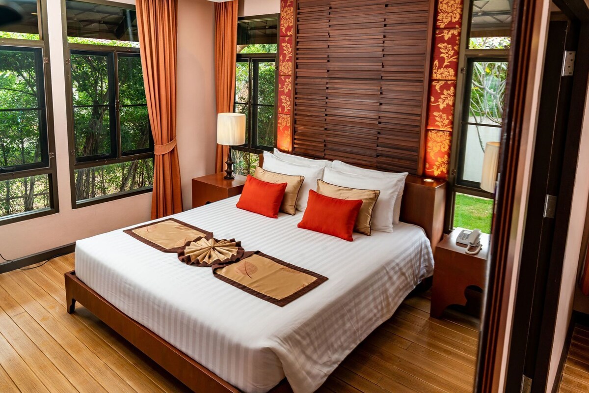 A hotel room with a large bed, wooden headboard, and a view of greenery through the windows.
