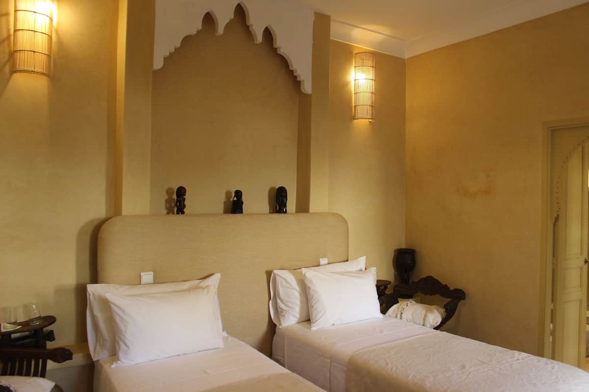 A hotel room with two beds, a large decorative headboard, wall-mounted lamps, and a small table with glasses.