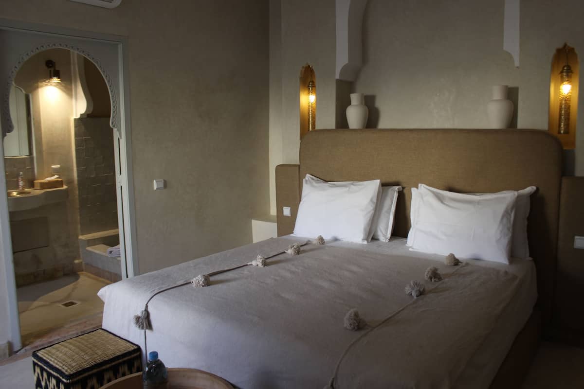 A bedroom with a large bed, a bedside table, a mirror, and a bathroom visible through an archway.
