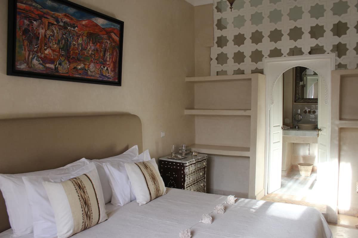 A bedroom with a bed, pillows, a nightstand, a painting, and a bathroom visible through an open door.