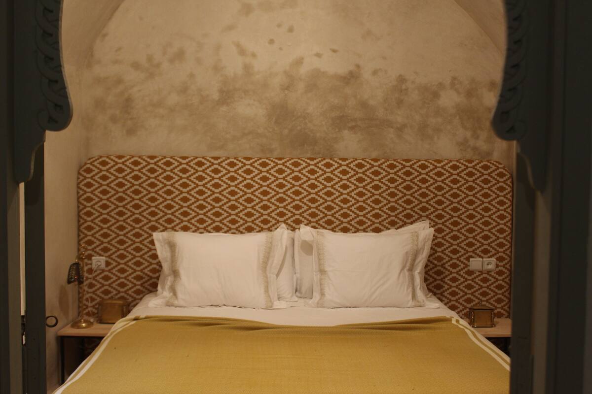A room with a patterned headboard, a bed with white linens, and a brown throw blanket.