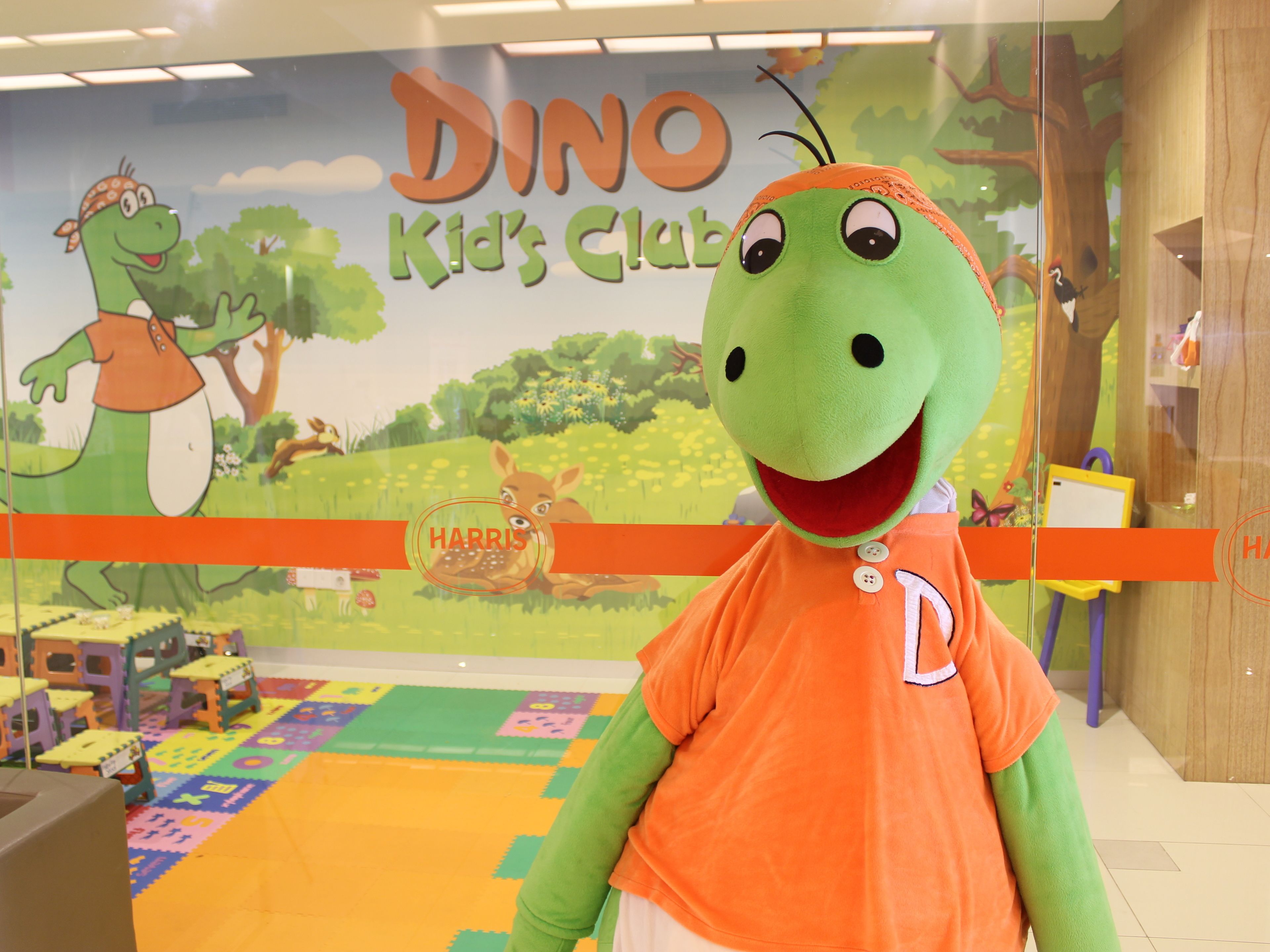 A dinosaur mascot in a children's play area with a mural of a dinosaur and a deer, labeled "DINO Kid's Club".