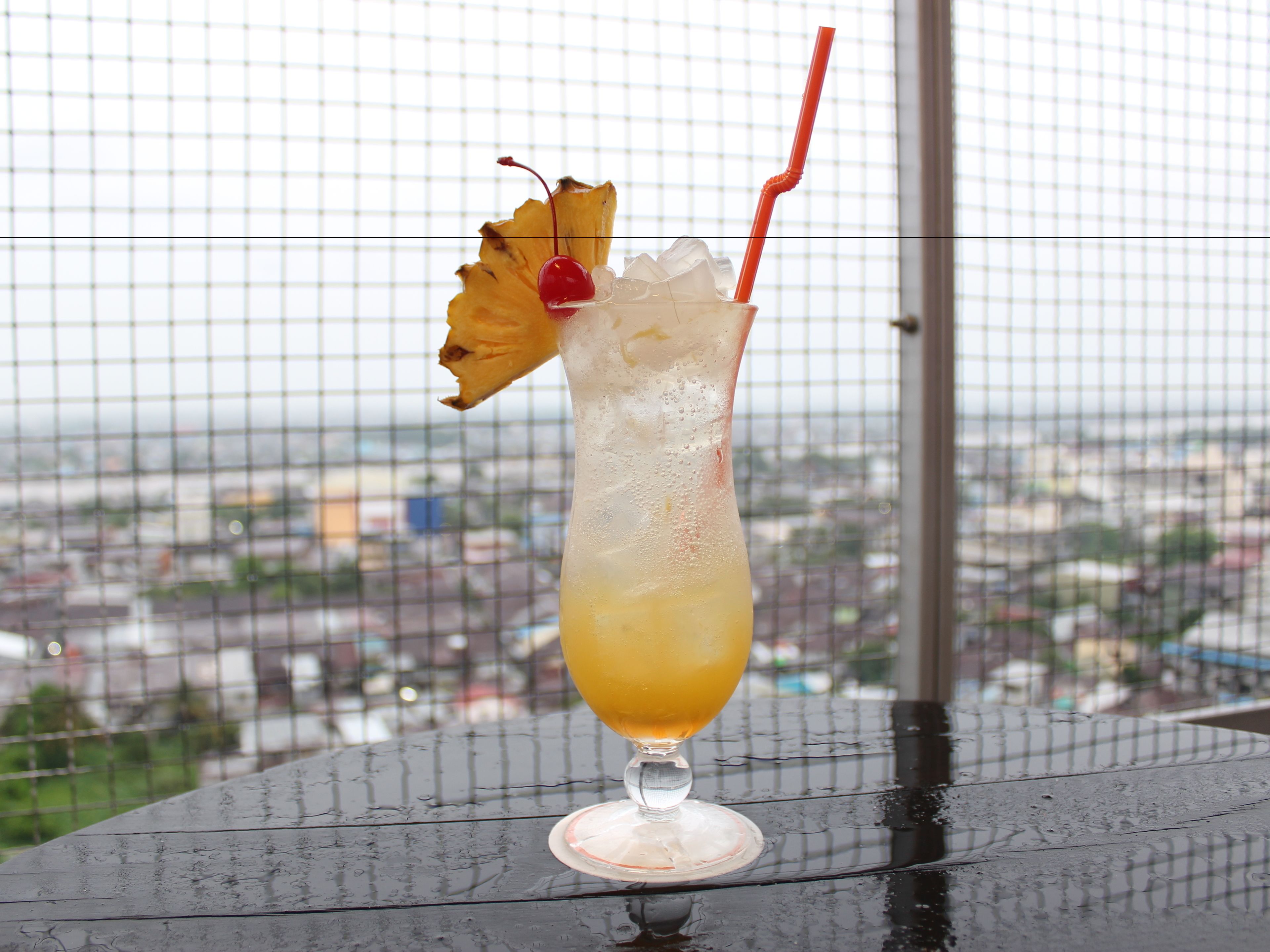 Un cocktail décoré d’un morceau d’ananas et muni d’une paille, posé sur une table avec une vue sur la ville en arrière-plan.