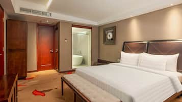 Une chambre d’hôtel avec un grand lit, un banc, une armoire en bois et une douche fermée par une paroi de verre.