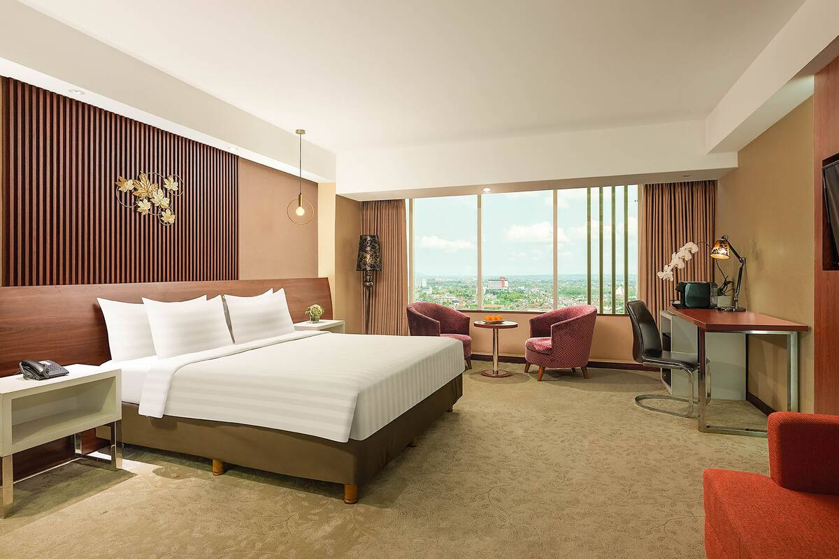 A hotel room with a large bed, a desk, two chairs, a small table with fruit, and a view of the city.