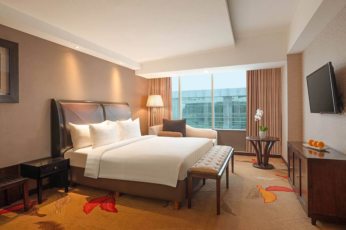 A hotel room with a large bed, a bedside table, a desk, a chair, a TV, and a view of the city.