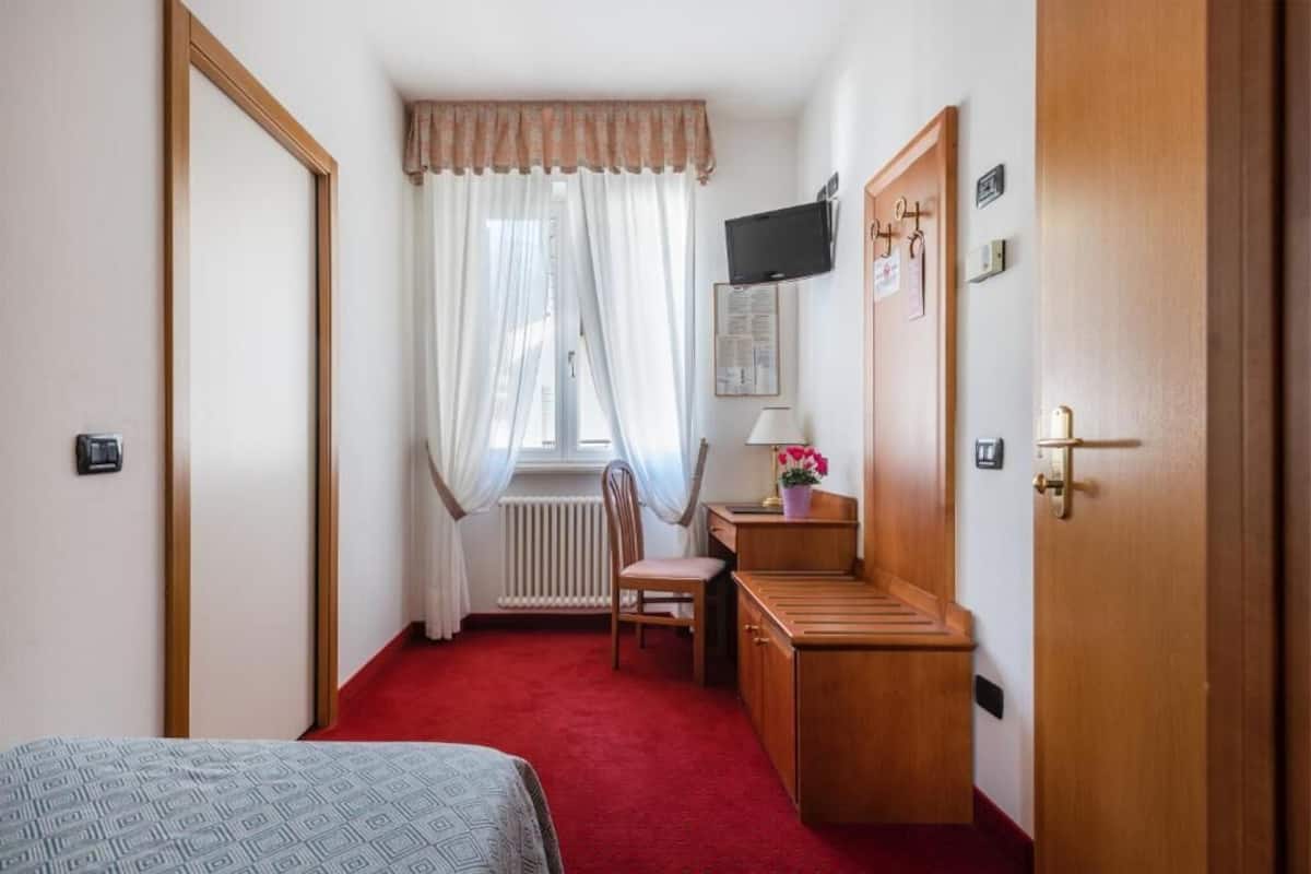 A hotel room with a bed, a desk with a chair, a radiator, a television mounted on the wall, and a door with a handle.