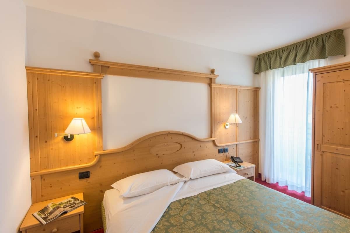 A hotel room with a wooden headboard, two bedside lamps, a telephone, a nightstand, and a window with curtains.