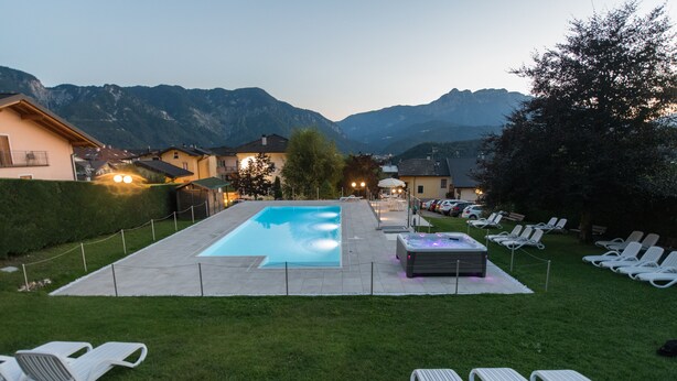A swimming pool with lounge chairs and a hot tub, surrounded by a fence and a view of mountains. - Hotel Lucia (Levico Terme)