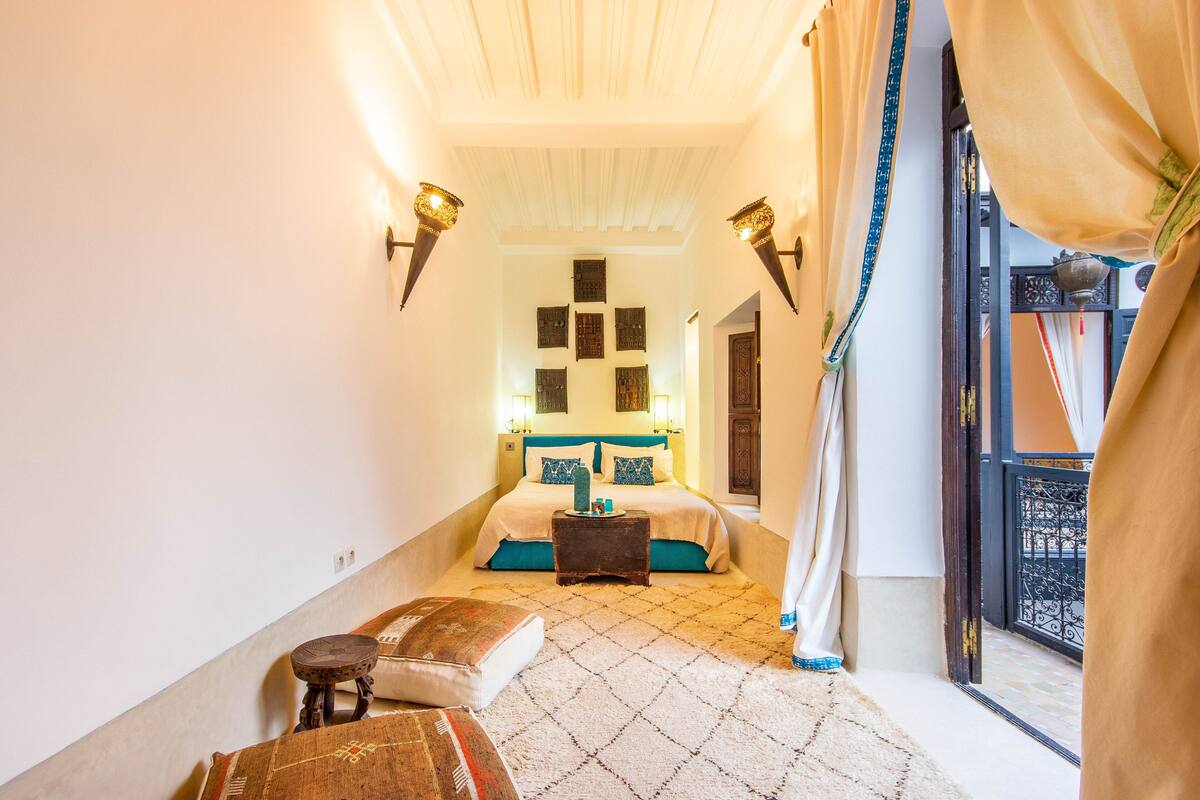 A bedroom with a bed, a small table, a chair, and a view of the outside balcony.