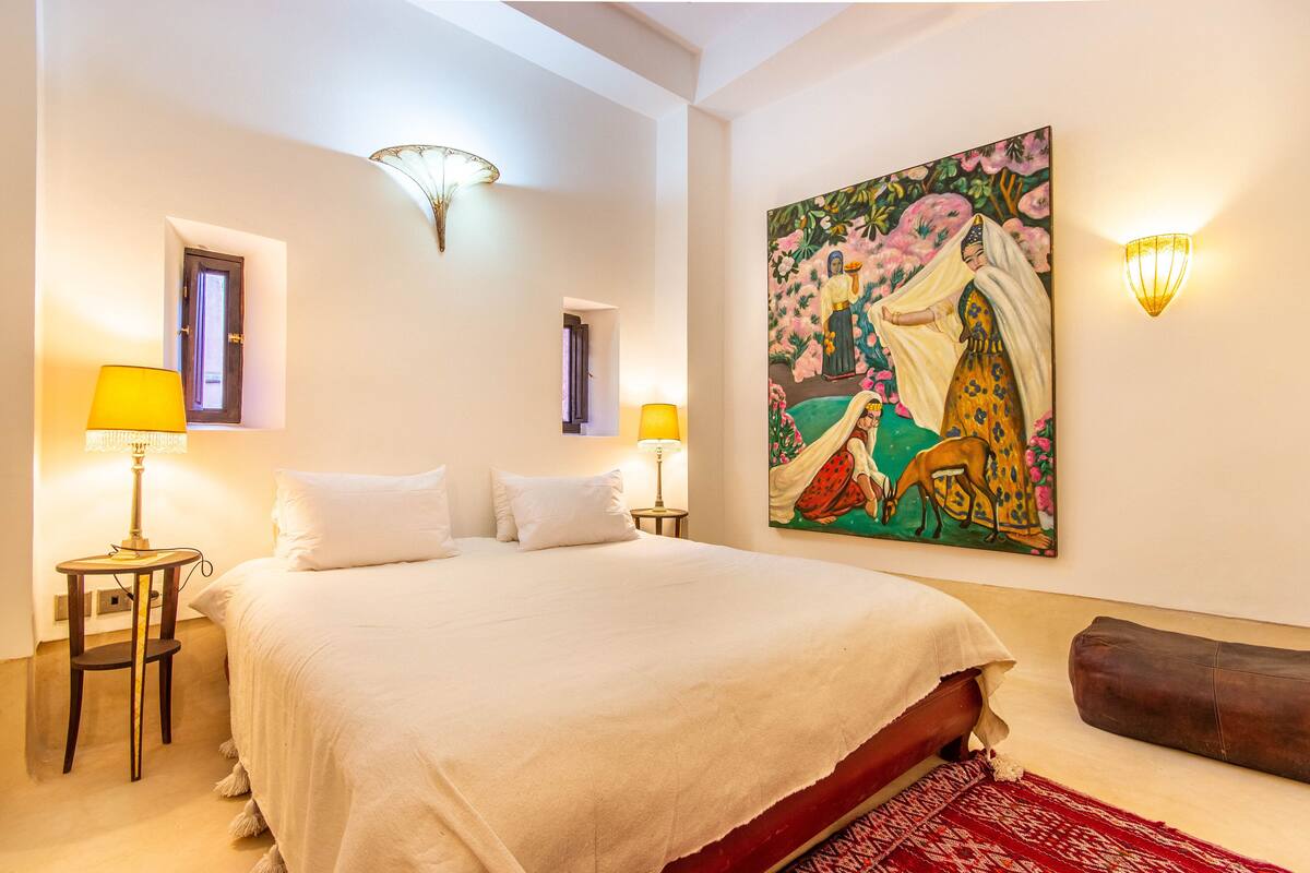 A spacious bedroom with a large bed, two bedside lamps, a red patterned rug, and a wall painting.