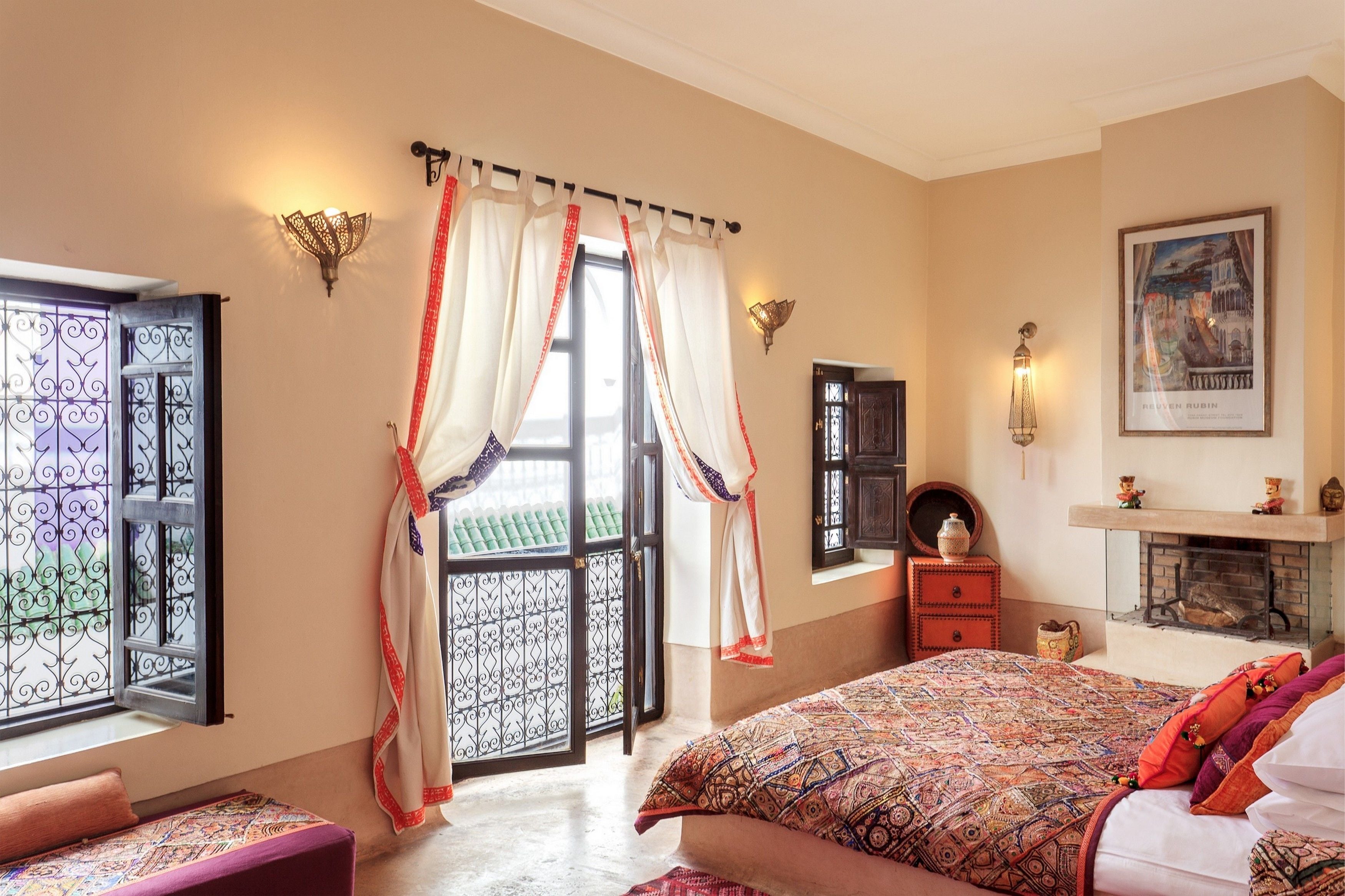 A bedroom with a large bed, a window with decorative curtains, a fireplace, and a wall-mounted shelf with decorative items.