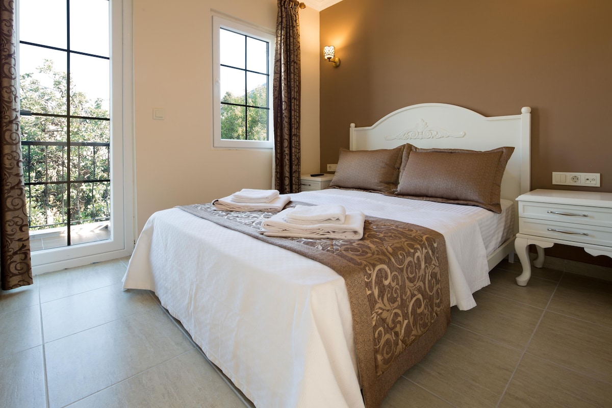 A neatly made bed with pillows and folded towels, a bedside table with a lamp, and a window with curtains.