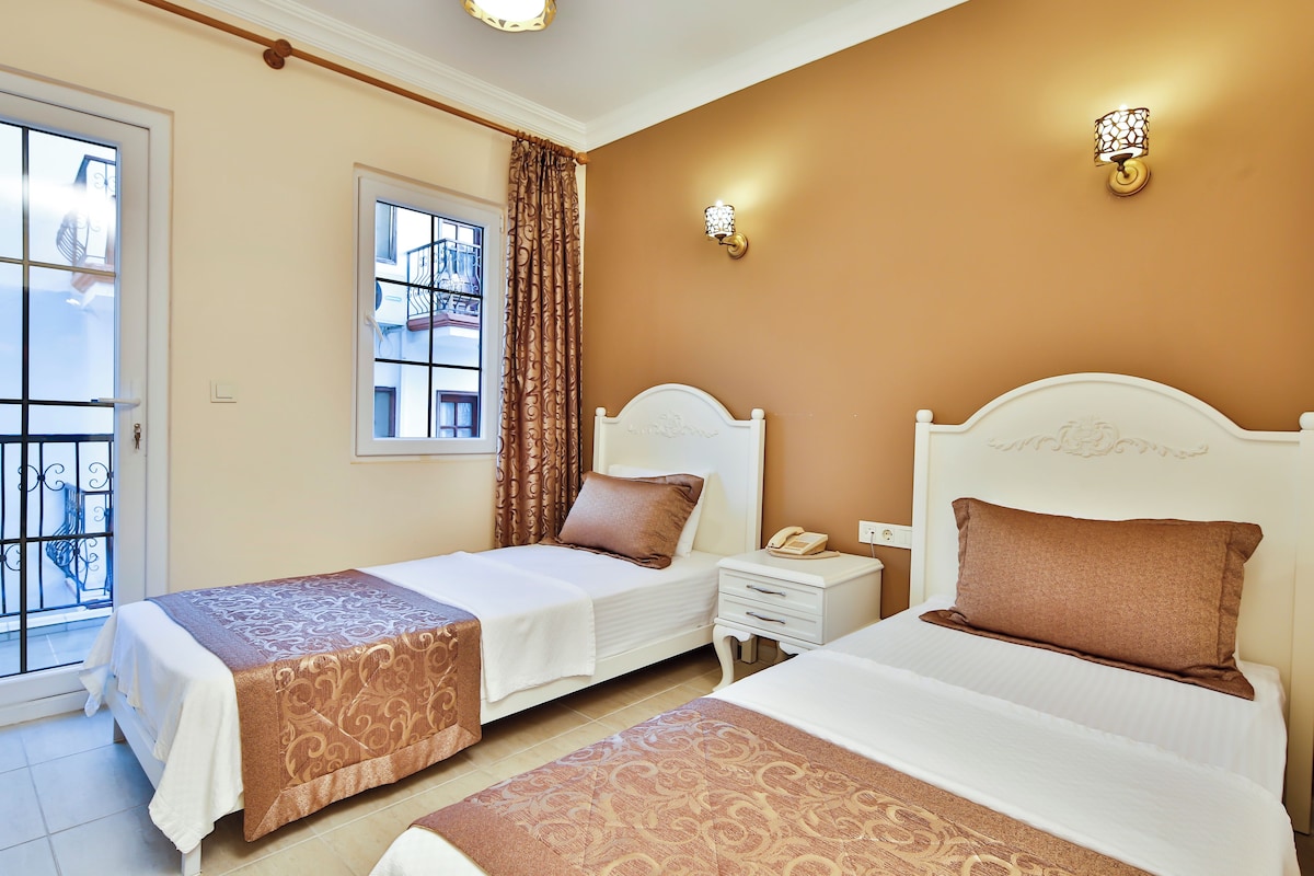 A hotel room with two beds, a window with curtains, a bedside table with a phone, and a wall-mounted light fixture.