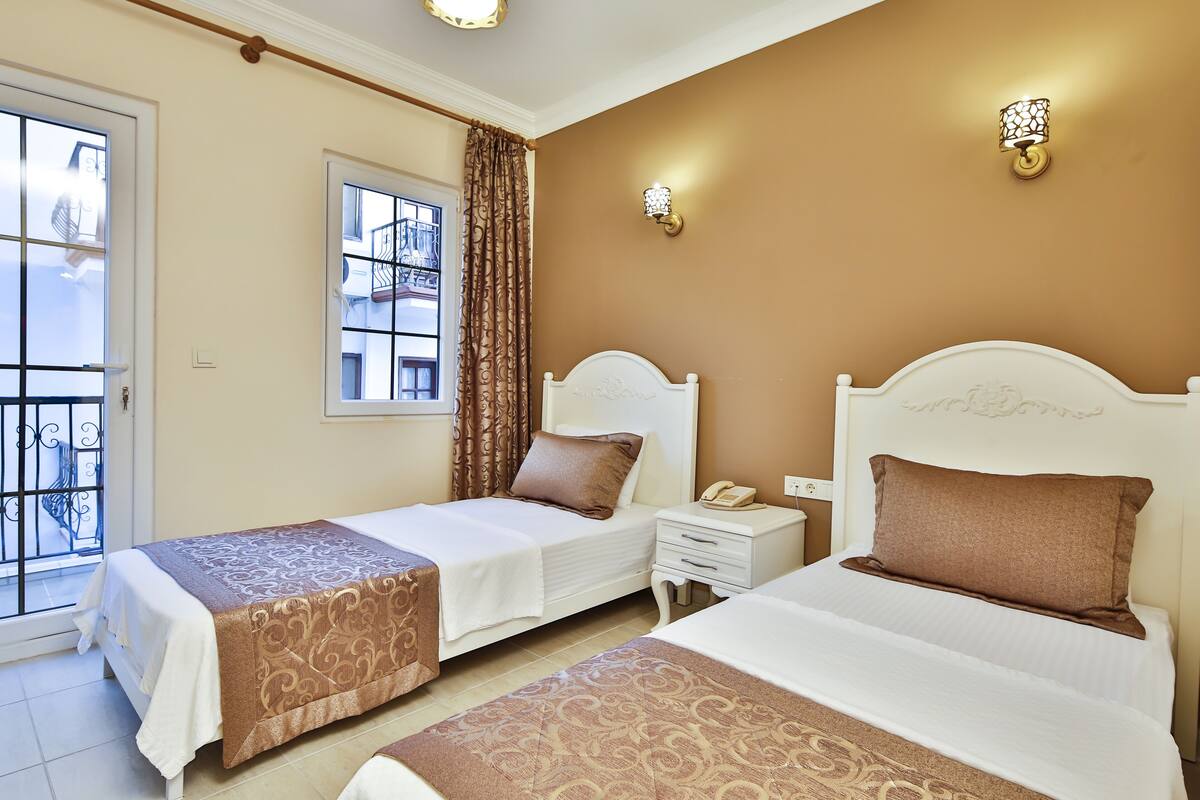 Habitación de hotel con dos camas, una ventana con cortinas, una mesita de noche con teléfono, y lámparas fijadas a la pared.