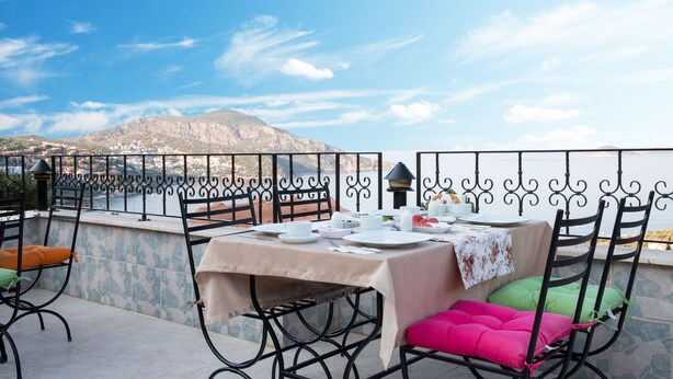 An outdoor dining area with a table set for two, surrounded by metal chairs with cushions, overlooking a scenic view of a body of water and a mountain. - Enda Boutique Hotel (Kas)