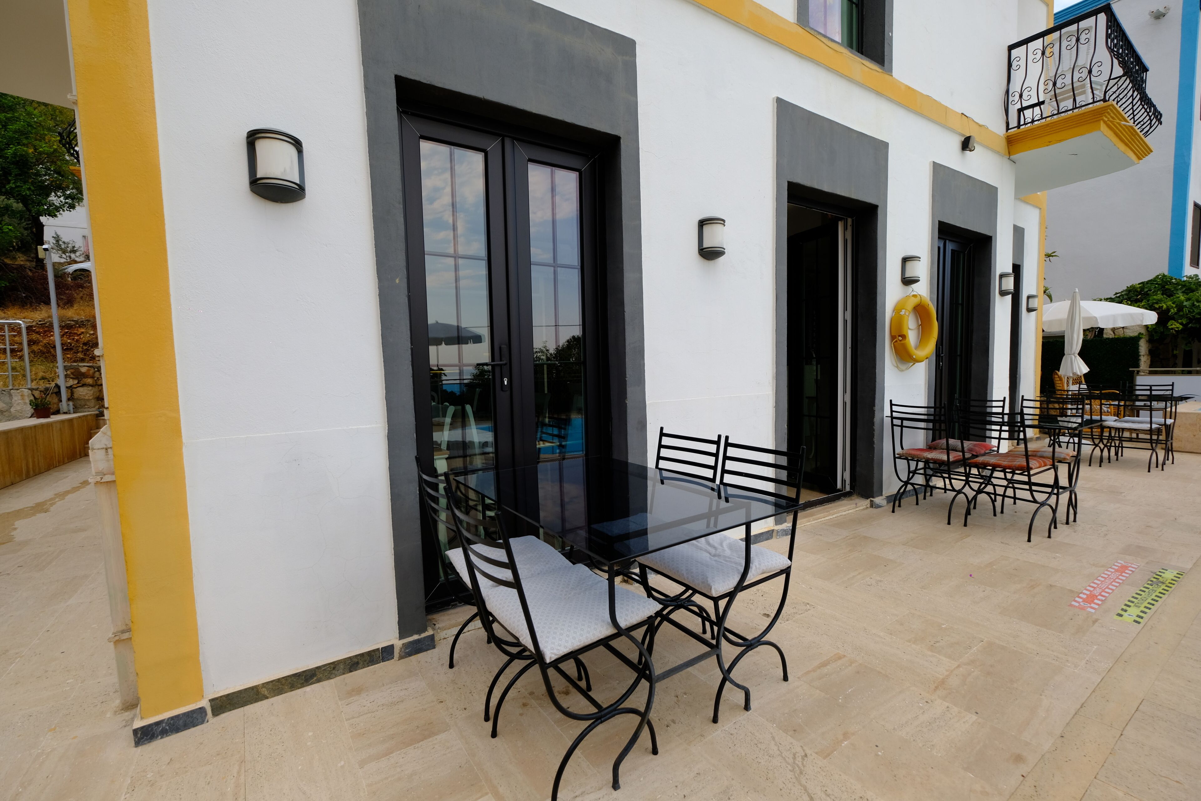 An outdoor patio with a glass table and metal chairs, a yellow pillar, and a black door.