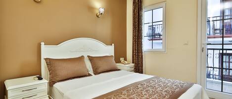 Habitación de hotel con cama blanca, almohadas marrones, mesita de noche, teléfono y ventana con vista al exterior.