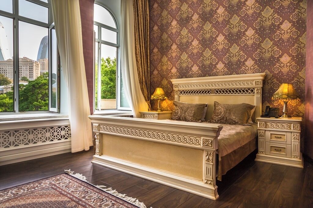 A bedroom with a large bed, ornate headboard, two bedside tables with lamps, a patterned wallpaper, and a view of buildings through a window.