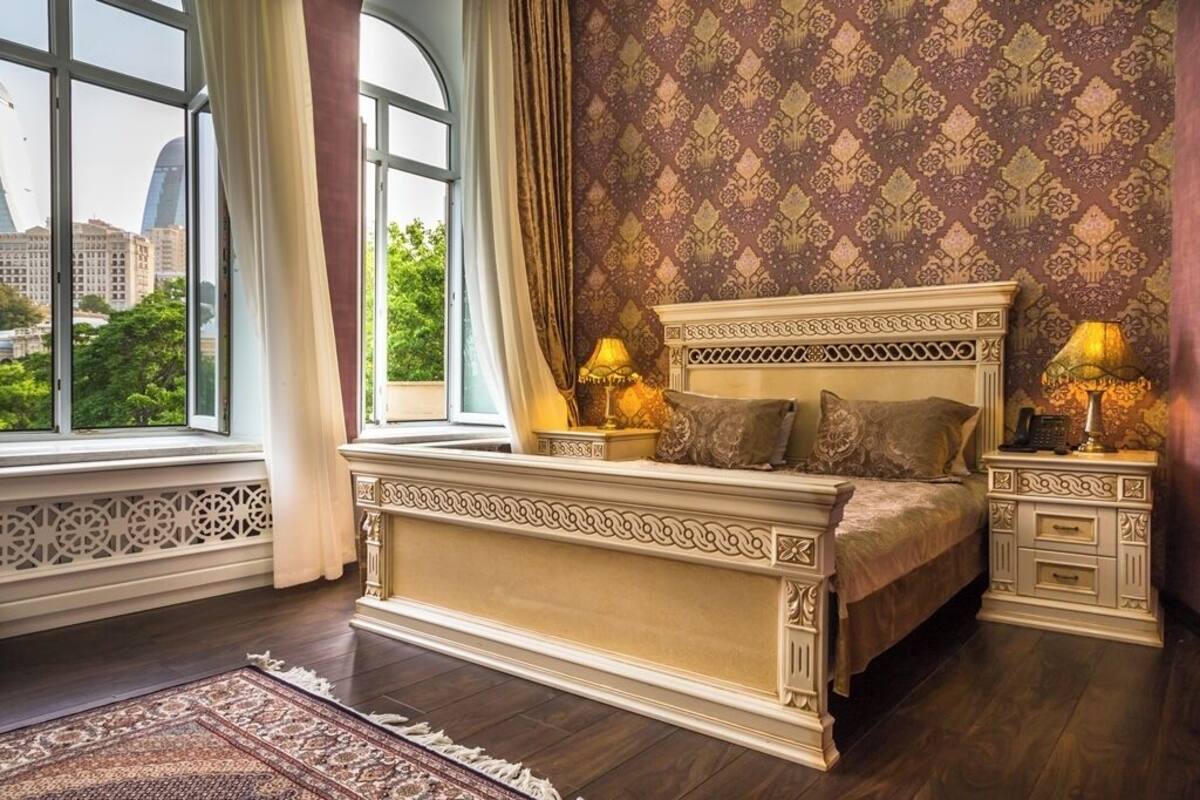 A bedroom with a large bed, ornate headboard, two bedside tables with lamps, a patterned wallpaper, and a view of buildings through a window.