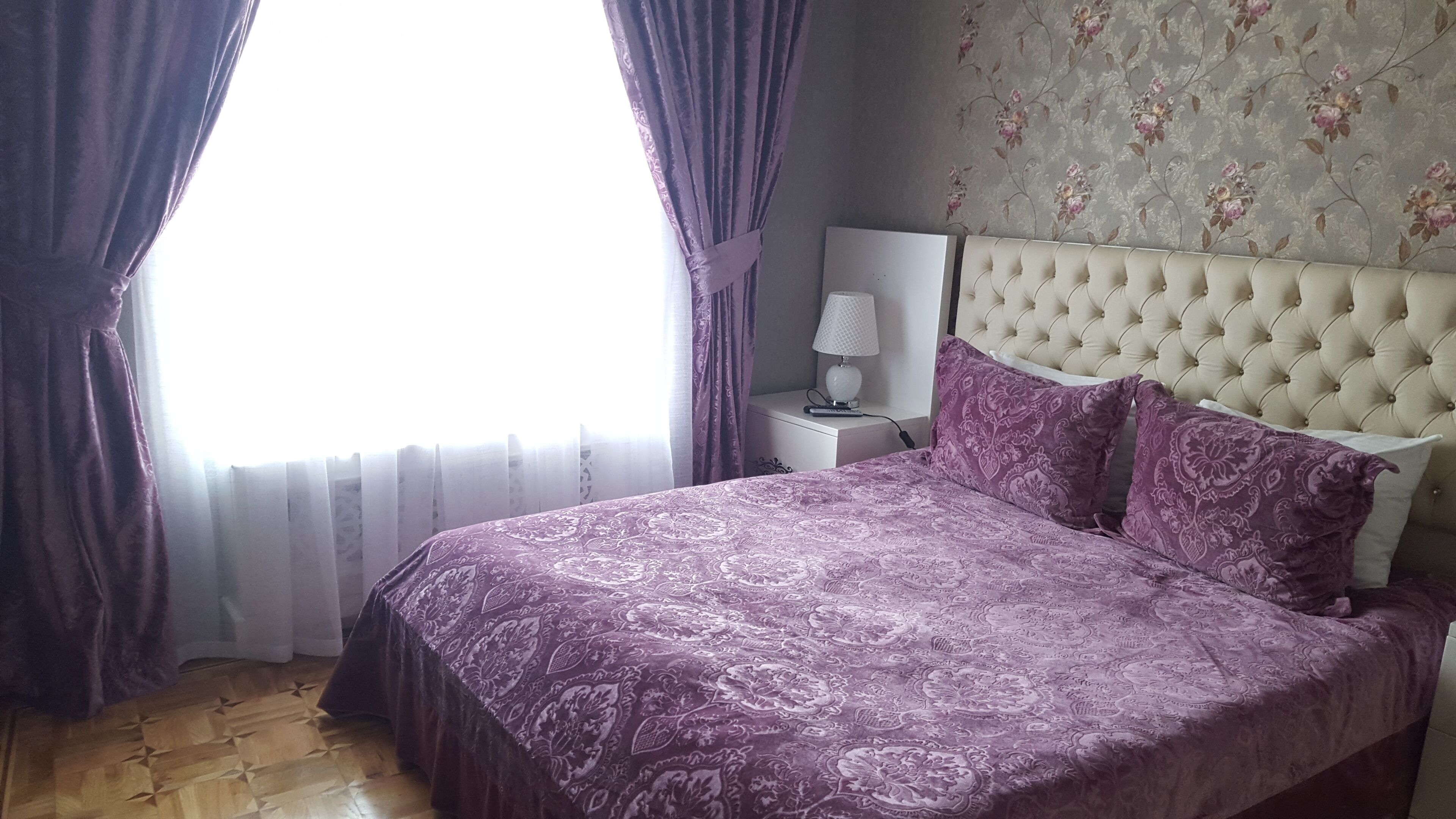A bedroom with a bed, curtains, a nightstand, a lamp, and a floral wallpaper.