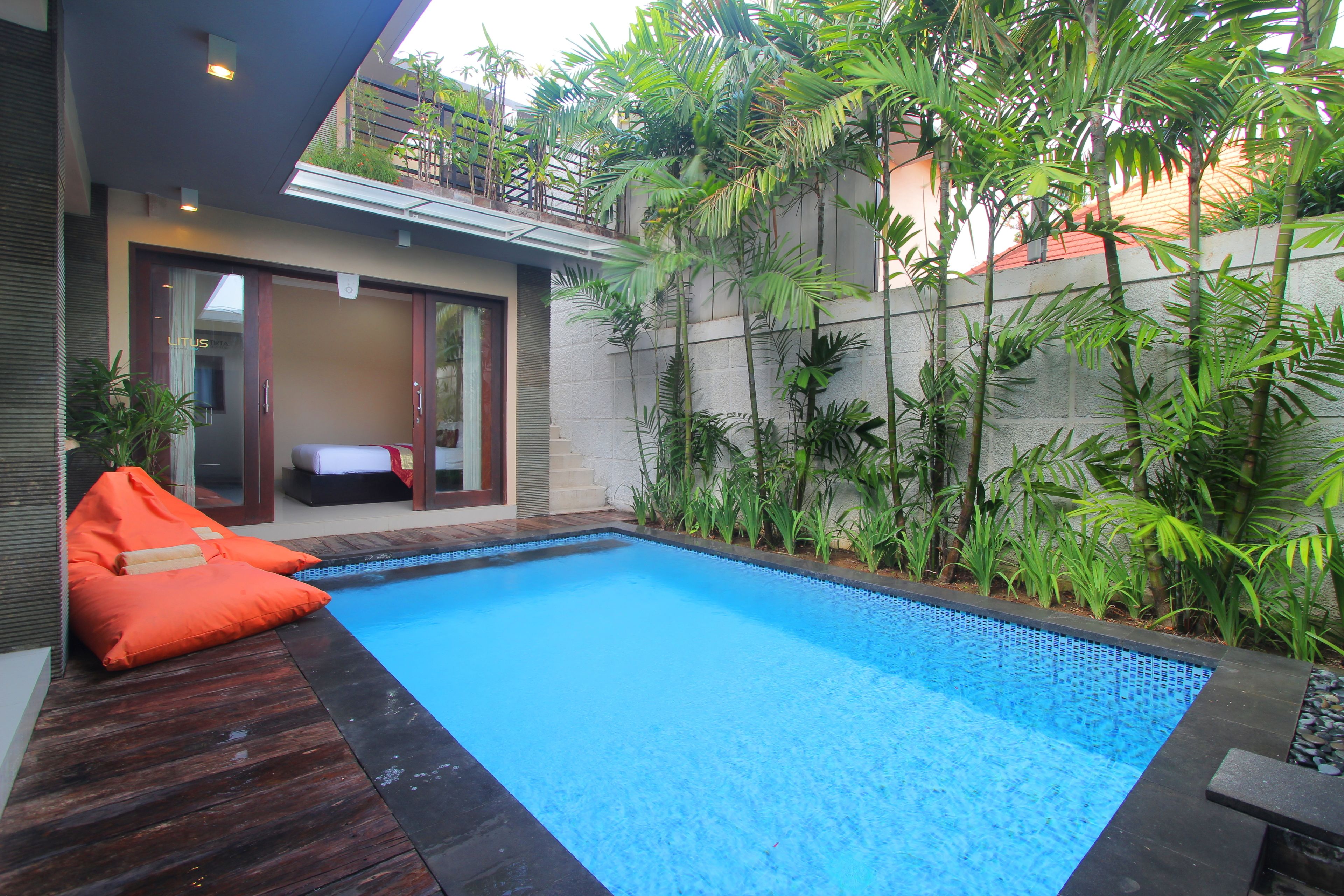 Une piscine dotée d’une terrasse en bois et de coussins orange, entourée d’une végétation luxuriante et bordée d’un bâtiment moderne aux portes vitrées.