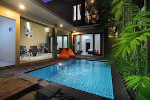 A modern indoor pool with a tiled floor, surrounded by wooden decking and a seating area with chairs and orange cushions. - Lotus Tirta Seminyak (Seminyak)