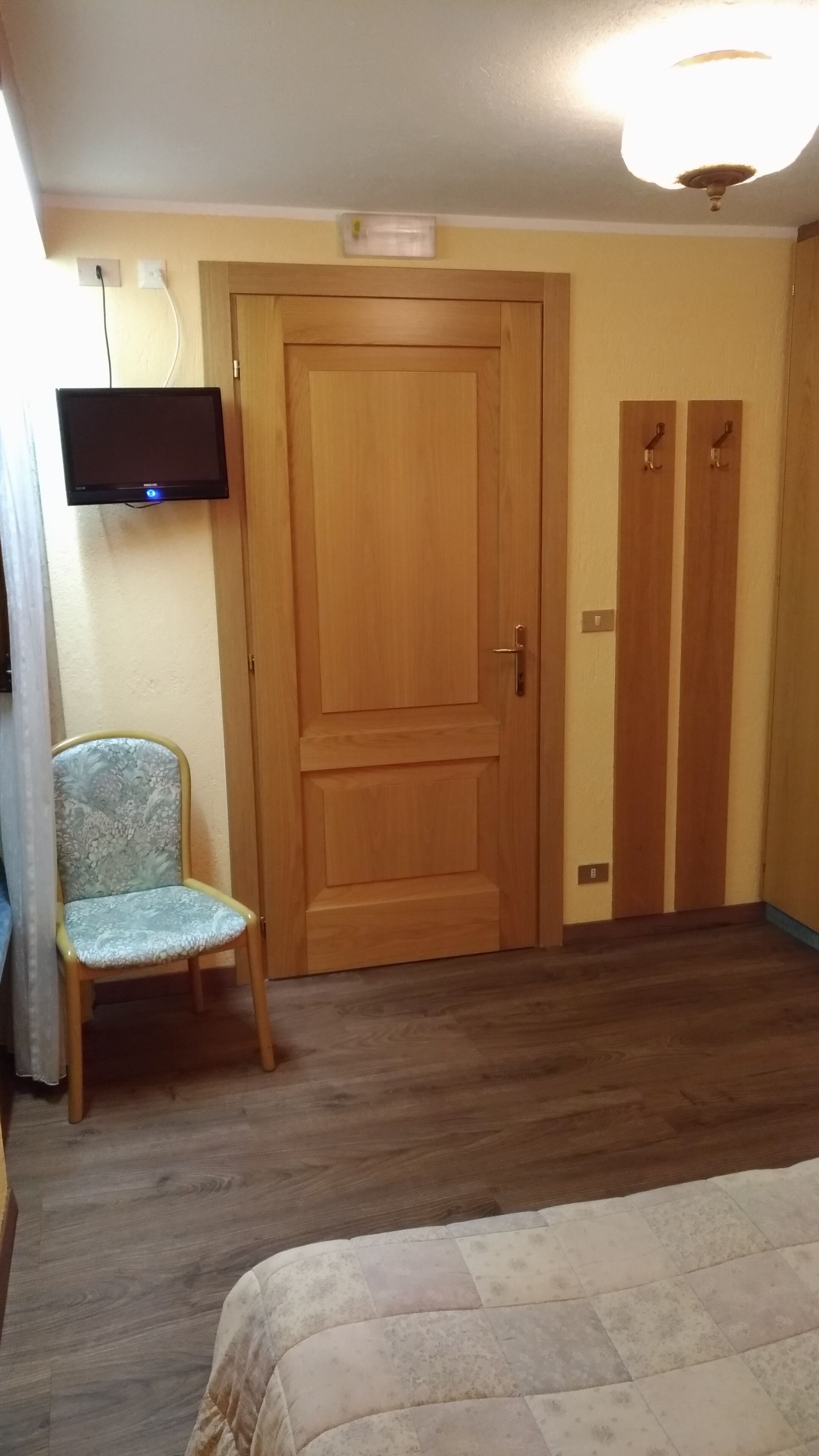 Un dormitorio con puerta de madera, una silla, televisor montado en la pared y una cama con colcha.