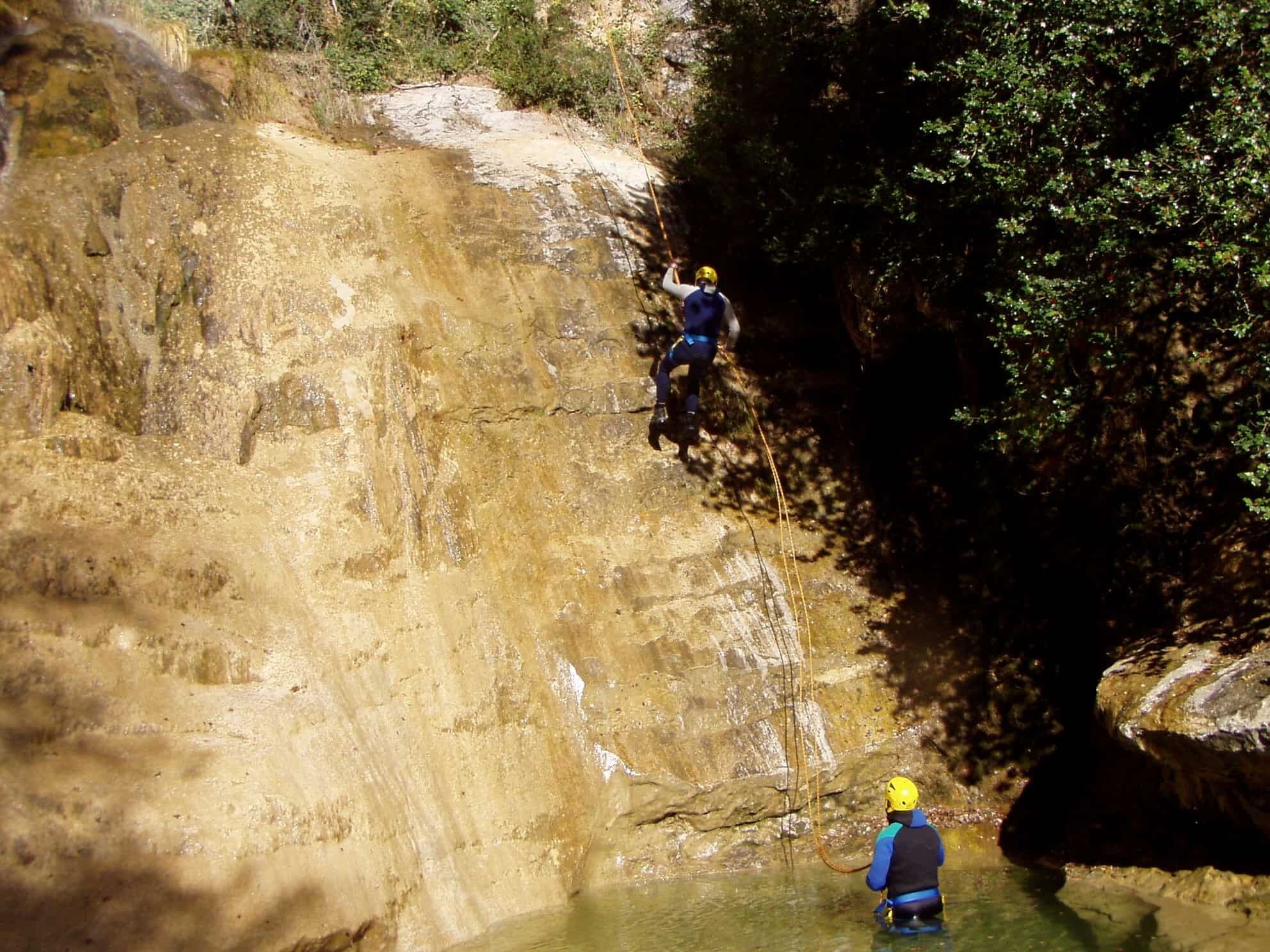 A person rappelling down a rocky cliff into a body of water, with another person standing in the water below.